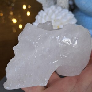 May include: A clear quartz crystal skull, carved with detailed features. The skull is held in a hand, highlighting its form. The crystal has a polished surface, reflecting light. This decorative item could be used for home decor or spiritual purposes.