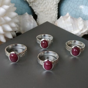 May include: Four silver rings, each with an oval, deep red gemstone. The rings feature a textured band design. They are displayed on a grey surface, with white seashells in the background.