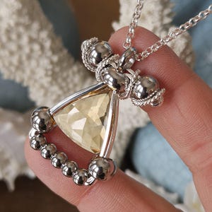 Peut inclure: Collier en argent orné d'un pendentif triangulaire en pierre jaune facettée. Le pendentif est encadré d'argent et orné de perles et de détails décoratifs argentés. Le collier est présenté sur un fond flou.