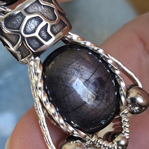 May include: Close-up of a silver pendant featuring a dark, oval-shaped gemstone. The gemstone is encased in a twisted silver wire design, with additional silver accents and a decorative top piece. The pendant is likely a piece of jewellery.