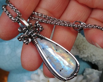 May include: A silver-toned necklace featuring a teardrop-shaped pendant with a rainbow moonstone. The pendant is intricately wire-wrapped and suspended from a delicate chain. The moonstone displays a play of colors, reflecting light. The necklace is a piece of jewelry.