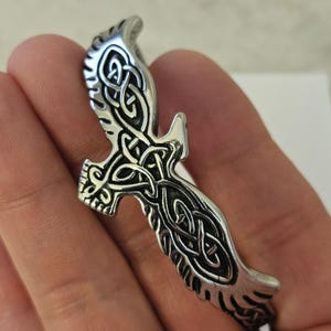 May include: A silver-toned metal cuff bracelet featuring a stylized bird design with intricate, embossed knotwork patterns across the wings and body. The surface has a polished finish with darkened recessed areas to highlight the detailed engravings.