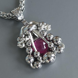 May include: A silver pendant necklace with a large, faceted, ruby-coloured gemstone. The pendant is encircled by small, shiny silver spheres and decorative silver elements. The chain is a thick, woven silver design.