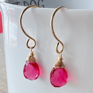 May include: A pair of gold earrings with pink teardrop-shaped gemstones. The gemstones are faceted and have a wire wrapped around the top.