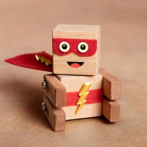 May include: A wooden toy robot wearing a red cape and a red mask with yellow eyes. The robot has a smiling face and a gold lightning bolt on its chest.