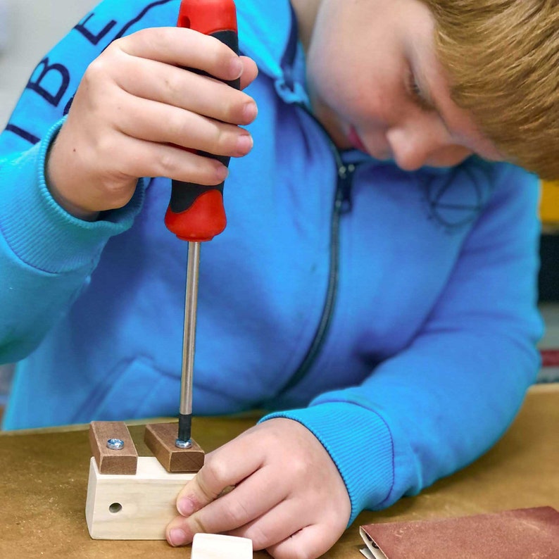 May include: A young person uses a red and silver screwdriver to assemble a wooden toy. The toy is made of light wood and has two small wooden blocks attached to it.
