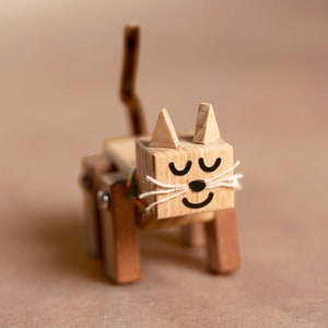 May include: A wooden toy cat with a painted face and white yarn whiskers. The cat has a closed-eye expression and a smiling mouth.