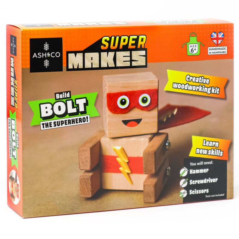 May include: A wooden toy superhero kit called "Build Bolt the Superhero!" from Super Makes by Ash & Co. The kit includes wooden pieces to build a superhero figure with a red cape, red mask, and a yellow lightning bolt on the chest. The box is orange and green with the text "Super Makes" and "Build Bolt the Superhero!" The box also includes instructions for building the superhero and a list of tools needed, including a hammer, screwdriver, and scissors.