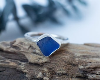 May include: A silver ring featuring a square, deep blue sea glass stone. The ring is set on a piece of driftwood, with a blurred background of foliage. The ring's design is simple and elegant.