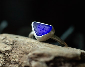 May include: A silver ring featuring a triangular piece of vibrant blue sea glass. The sea glass is set in a silver bezel, creating a striking contrast. The ring is displayed on a piece of driftwood against a dark background.