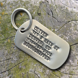 May include: A silver metal pet ID tag with a ring. The tag has stamped text that reads "OLIVER, 345 W MAIN ST, DENVER, CO, 234-567-8901, MICROCHIPPED". The tag is on a weathered wood surface.