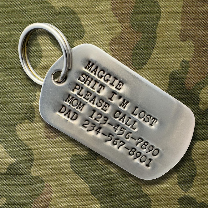 May include: A silver dog tag with a ring, featuring stamped text. The text reads "MAGGIE SHIT I'M LOST PLEASE CALL MOM 123-456-7890 DAD 234-567-8901". The background is camouflage fabric.