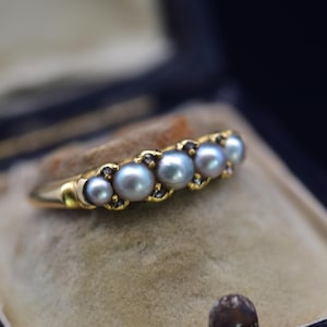 May include: A gold ring featuring five gray pearls. The pearls are set in a row, with small accent stones between each pearl. The ring is displayed on a beige surface, with a dark background.