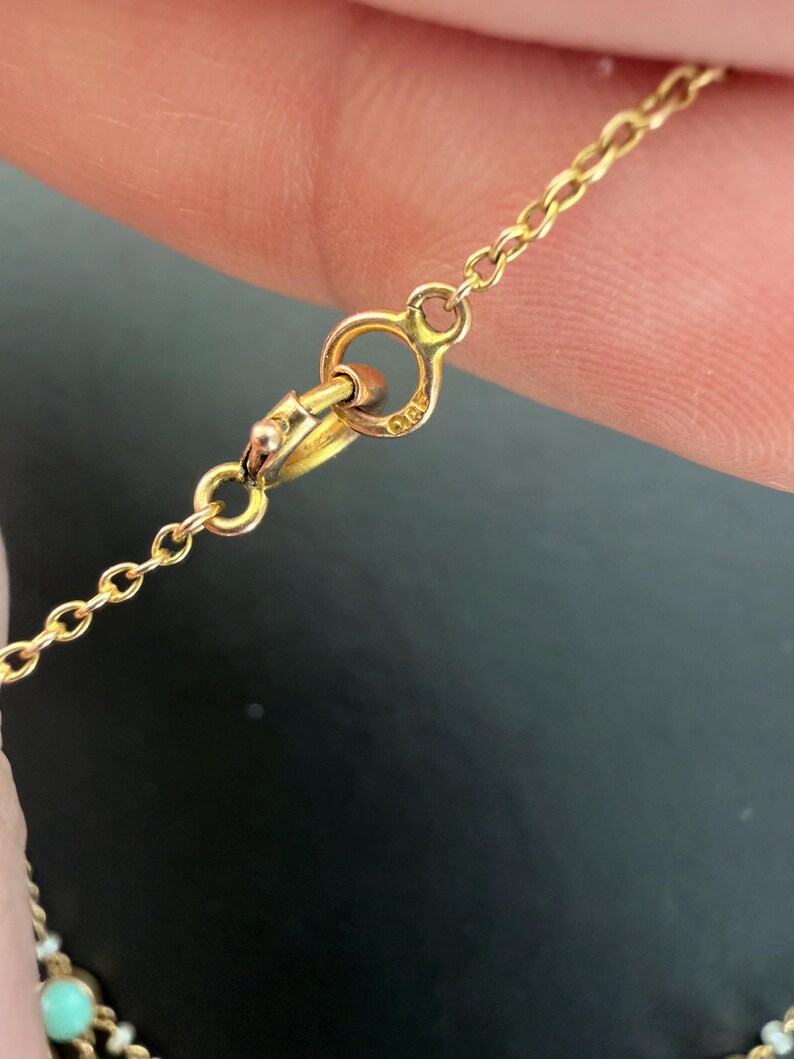 May include: Close-up of a gold necklace clasp and chain. The chain is delicate, with small links. The clasp is a simple design, with a small loop for attaching the necklace. The necklace has small turquoise and white beads.