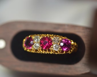 Könnte beinhalten: Ein goldener Ring mit fünf Edelsteinen: drei roten Rubinen und zwei Diamanten. Der Ring ist in einem strukturierten Goldband gefasst und wird in einer Holzschatulle präsentiert. Die Rubine sind tiefrot. Ein Schmuckstück.