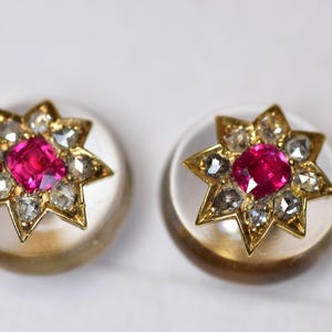 Puede incluir: Un par de pendientes en forma de estrella. Cada pendiente presenta una gema rosa vibrante en el centro, rodeada de gemas claras más pequeñas y engastada en metal dorado. Los pendientes descansan sobre una base circular blanca.