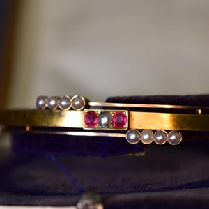 May include: A gold bangle bracelet featuring a central rectangular setting with two vibrant pink gemstones flanking a single white pearl. The bracelet is accented by two parallel rows of four small white pearls, positioned above and below the center.