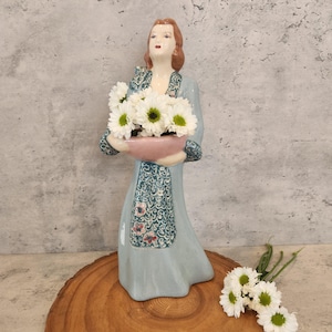 May include: A ceramic figurine of a woman in a blue dress with a floral pattern, holding a pink planter with white flowers. The figurine is standing on a wooden surface.