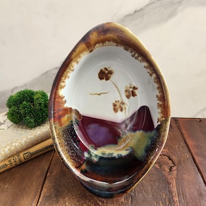 May include: Oval-shaped ceramic bowl with a white interior featuring brown floral designs and a purple and green base. The rim is a mix of brown and orange hues, creating a rustic aesthetic. The bowl is likely handmade.