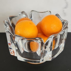 Könnte beinhalten: Eine klare Glasschüssel mit einem einzigartigen, facettierten Design enthält eine Gruppe von vier leuchtend orangefarbenen Mandarinen. Die Schüssel hat eine breite, offene Oberseite und einen abgerundeten Boden.