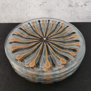 May include: A stack of clear glass coasters with a radial design. The design features alternating black, gold, and gray teardrop shapes radiating from a central circle. The coasters are stacked on a dark surface.