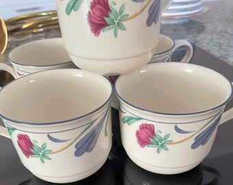 May include: A set of white ceramic teacups with a floral design. Each cup features a magenta flower and blue and green leaf accents, with a blue rim. The cups are stacked and arranged on a dark surface.