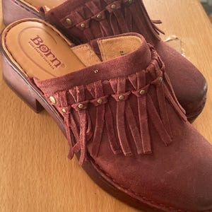 May include: Burgundy suede clogs with fringe detailing and brass-colored studs. The clogs have a wooden sole and the brand name "Born" is visible on the insole. These shoes are a stylish addition to any wardrobe.