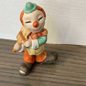 May include: A ceramic clown figurine with a green hat, orange hair, and a red nose. The clown is holding a yellow and orange musical instrument.