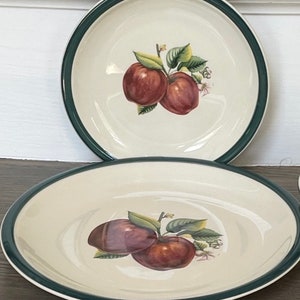 Puede incluir: Dos platos de cerámica blanquecina con borde verde. Cada plato presenta una ilustración detallada de manzanas rojas con hojas verdes. Los platos son redondos y ovalados, adecuados para servir alimentos.