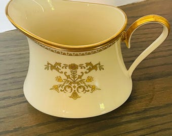 Puede incluir: Cremera de color crema con borde y asa dorados. La cremera presenta un diseño floral en tonos dorados y marrones. El asa es curvada y el borde tiene un borde decorativo. Una pieza clásica para servir crema o leche.