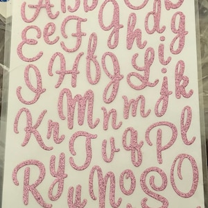 May include: Sheet of pink glitter alphabet stickers. The stickers include uppercase and lowercase letters, from A to Z. The package is labeled "sticko" and includes the text "STICKERS AUTOCOLLANTS ETIQUETAS".
