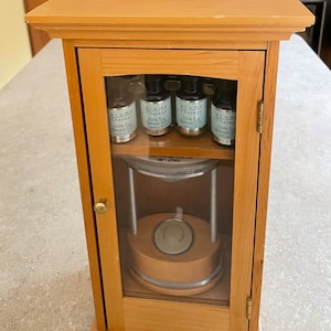 Puede incluir: Armario difusor de aceites esenciales Aroma Therapy de madera clara. El armario tiene una puerta de cristal y contiene cuatro pequeñas botellas de aceites esenciales y un difusor. Las botellas están etiquetadas con los nombres de los aceites esenciales.