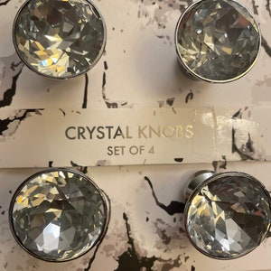 Può includere: Set di quattro pomelli di cristallo con finitura argentata. I pomelli sono rotondi e hanno un design sfaccettato. La confezione ha uno sfondo marmoreo con il testo "CRYSTAL KNOBS SET OF 4".