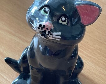 May include: A black ceramic cat figurine with pink ears, white whiskers, and green eyes. The cat is sitting with its head tilted up.