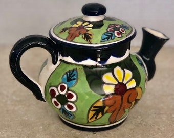 May include: A small green ceramic teapot with a blue handle and lid. The teapot is decorated with colorful flowers and leaves.