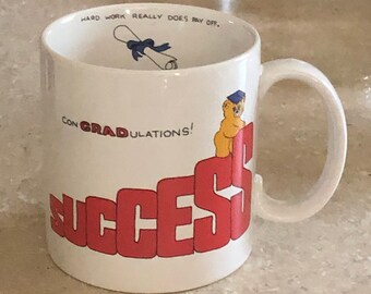 May include: White ceramic mug with the words "CON GRADULATIONS!" and "SUCCESS" in red block letters. A cartoon figure in a graduation cap is perched on the "S". Inside the mug, a rolled diploma with a blue ribbon is visible. The text "HARD WORK REALLY DOES PAY OFF" is also printed on the mug.
