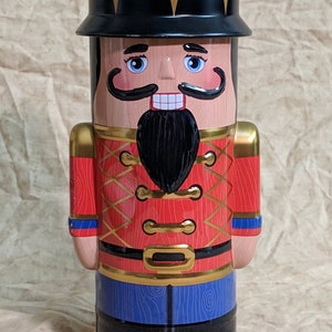 May include: A decorative tin shaped like a nutcracker soldier. The tin features a black hat with gold accents, a painted face, and a red uniform with gold trim. The bottom portion is blue with black boots.