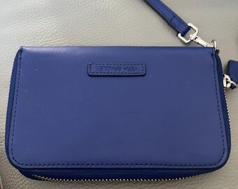 Può includere: Un portafoglio in pelle blu con chiusura a zip e logo Vera Bradley. Il portafoglio ha un cinturino da polso.