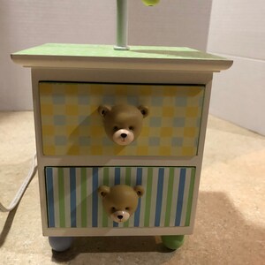 May include: A small, white nightstand with two drawers, each adorned with a bear-shaped knob. The drawers feature a checkered pattern in yellow and blue, and a striped pattern in blue and green. The top is light green.