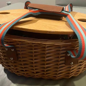 May include: A woven wicker picnic basket with a wooden lid. The lid has two circular cutouts and a brown leather handle. The basket has a striped blue, red, and white strap for carrying.