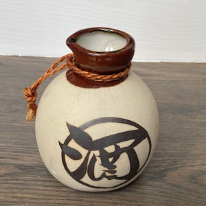 May include: A small, white ceramic sake bottle with a brown lid and a brown rope tied around the neck. The bottle has a black and white design of a Japanese character on the front.