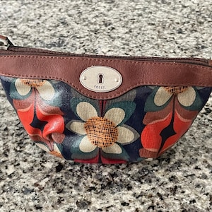 May include: A small, floral-patterned pouch with a brown leather trim and a zipper closure. The pouch features a navy blue background with large, colorful flower designs in red, white, and teal. The Fossil logo is visible.