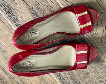 May include: A pair of red patent leather ballet flats with bow accents. The shoes have a glossy finish and feature a gold-colored bar on the bow. The brand name "...me Too" is visible inside the shoe.
