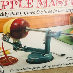 Könnte beinhalten: Ein teal Apfelschäler mit einem roten Apfel, der geschält wird. Der Schäler hat eine Kurbel und einen Saugfuß. Der Text "APPLE MASTER" und "Quickly Pares, Cores & Slices in one easy" sind sichtbar.