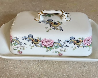 Könnte beinhalten: Eine weiße Porzellan-Butterdose mit Blumenmuster. Der Deckel hat einen goldenen Griff und ein Blumenmuster. Die Schale hat einen gewellten Rand.