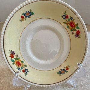 May include: A vintage, cream-colored plate with a floral design. The plate features a white center, a cream-colored outer ring, and a gold trim. The rim has a beaded edge. Colorful floral clusters are arranged around the plate's edge.