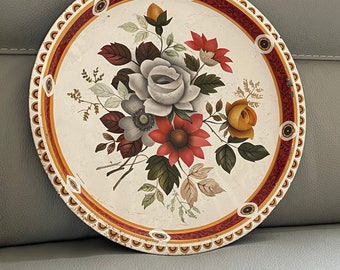 Può includere: Vassoio rotondo vintage con un design floreale. Il vassoio ha uno sfondo bianco e un bordo rosso e oro. La composizione floreale comprende rose, margherite e altri fiori nei toni del rosso, bianco e giallo.