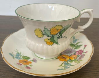 May include: A vintage teacup and saucer set. The white teacup has a green rim and handle, with yellow and green floral designs. The matching saucer has a scalloped edge and gold trim, with similar floral patterns.