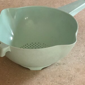 May include: A light green plastic colander with a handle and a pouring spout. The colander has a bowl shape with small holes in the bottom for draining. It has a handle on one side and a spout on the other.