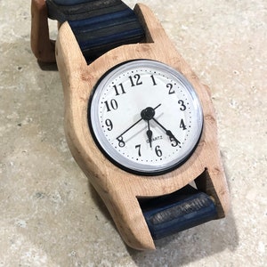May include: A wooden wristwatch with a light-colored wood frame and a white clock face. The watch band is made of dark blue and black layered wood. The clock face has black numerals and hands, and the word "QUARTZ" is printed at the bottom.